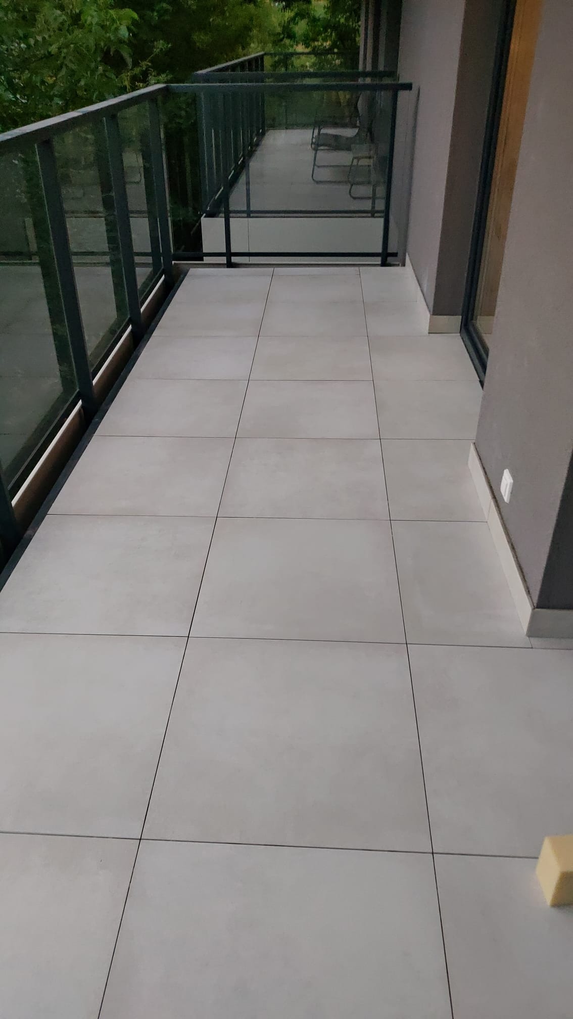 Wyłożony jasnymi płytkami taras z szklaną balustradą i widokiem na zieleń, z fragmentem żółtej gąbki w rogu kadru.