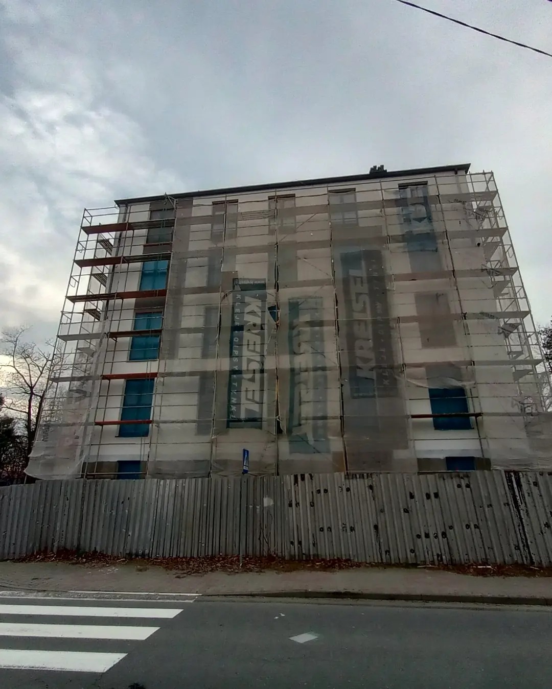 Wielopiętrowy budynek w Bystrem w trakcie prac remontowych, osłonięty rusztowaniami i siatką z reklamą producenta materiałów budowlanych, widok z dołu z poziomu ulicy.