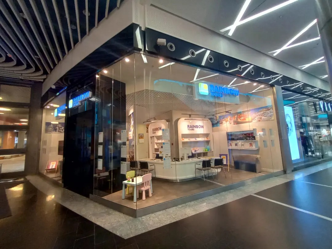 Przeszklony lokal biura podróży Rainbow Tours w centrum handlowym, widoczne wnętrze z biurkami i dekoracjami podróżniczymi, mały stolik z krzesełkami dla dzieci przy wejściu.