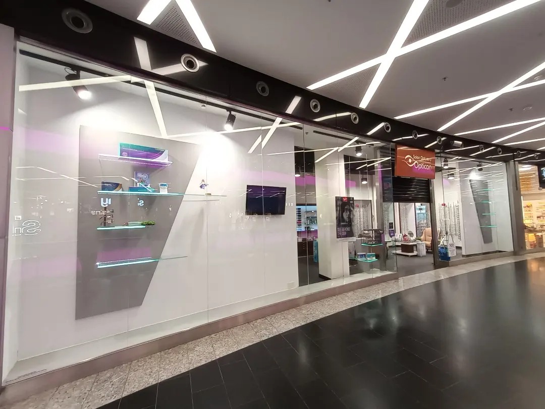 Przeszklona fasada salonu optycznego w centrum handlowym, widoczne półki z produktami i logo firmy.