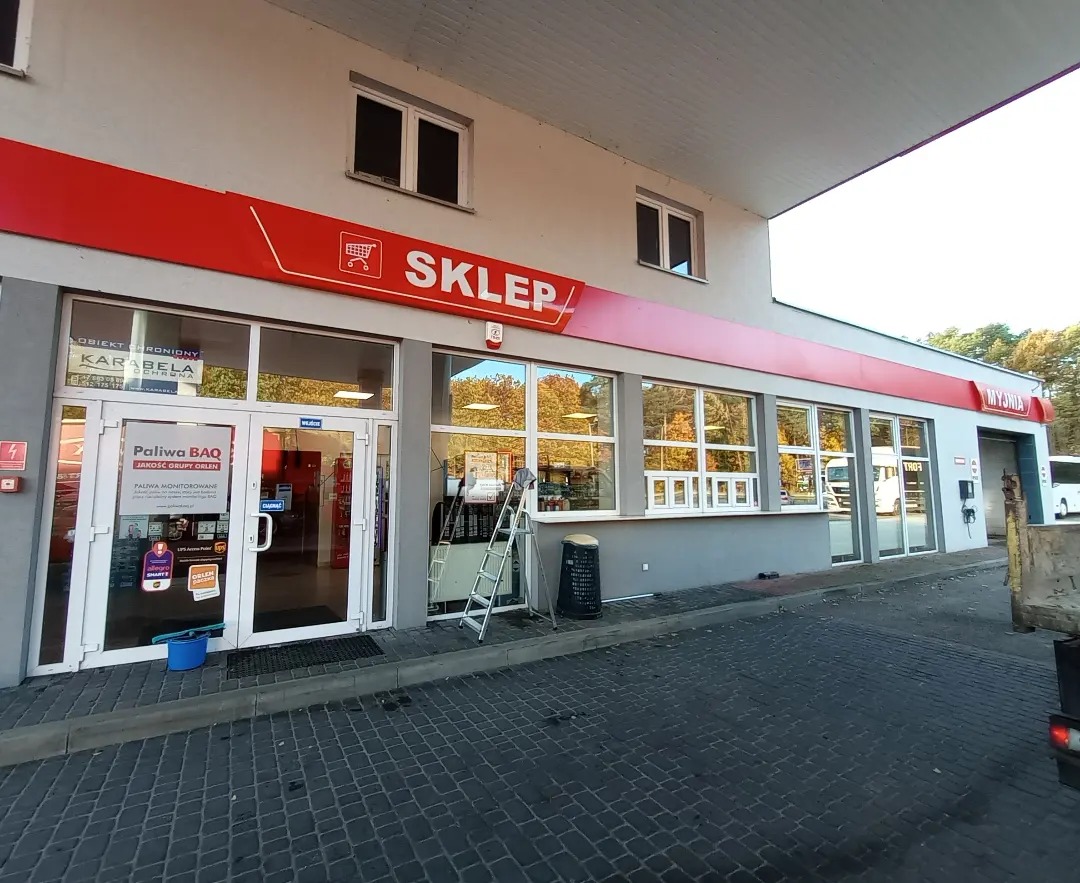 Zewnętrzny widok sklepu na stacji benzynowej z widoczną drabiną opartą o okno, sugerującą prace porządkowe. Na chodniku widoczne wiadro i mop.
