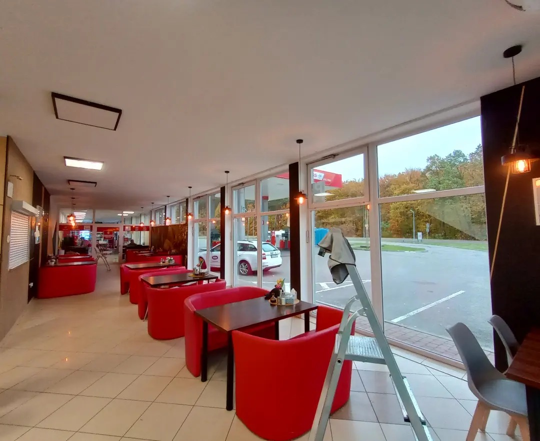 Wnętrze lokalu z czerwonymi sofami i stolikami, widok na stację benzynową przez duże okna, drabina oparta o okno sugerująca prace porządkowe.