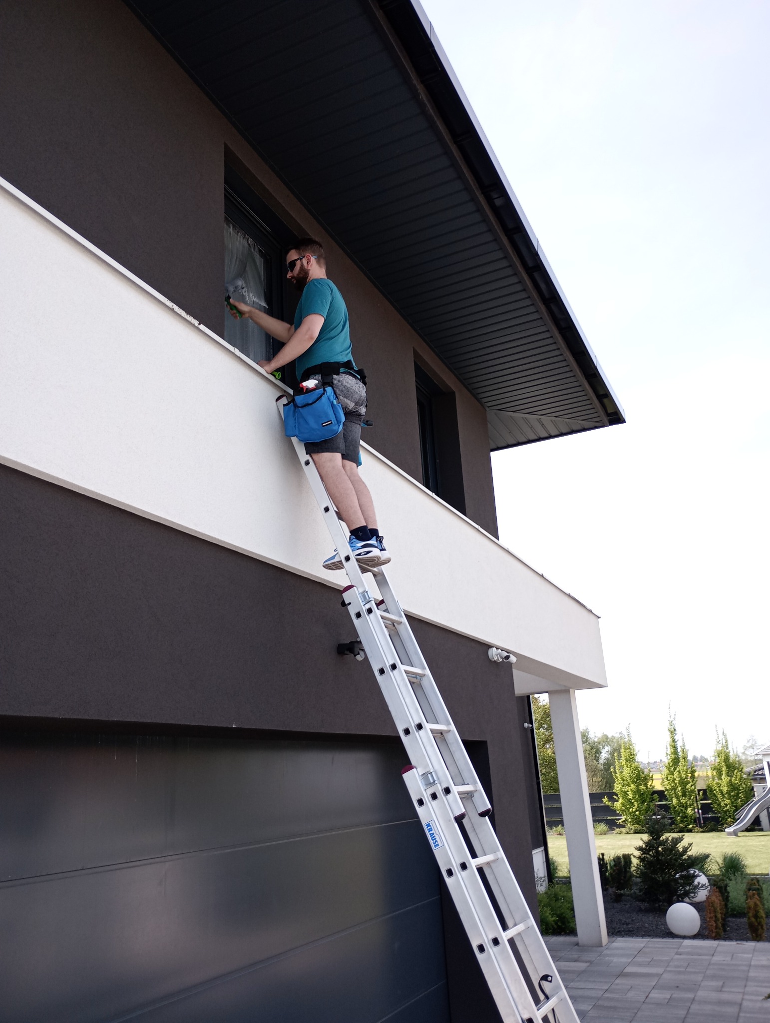 Pracownik na drabinie czyści okno na wysokości w nowoczesnym domu z ciemną elewacją i białymi akcentami architektonicznymi. Widoczny pas narzędziowy i sprzęt do mycia okien.