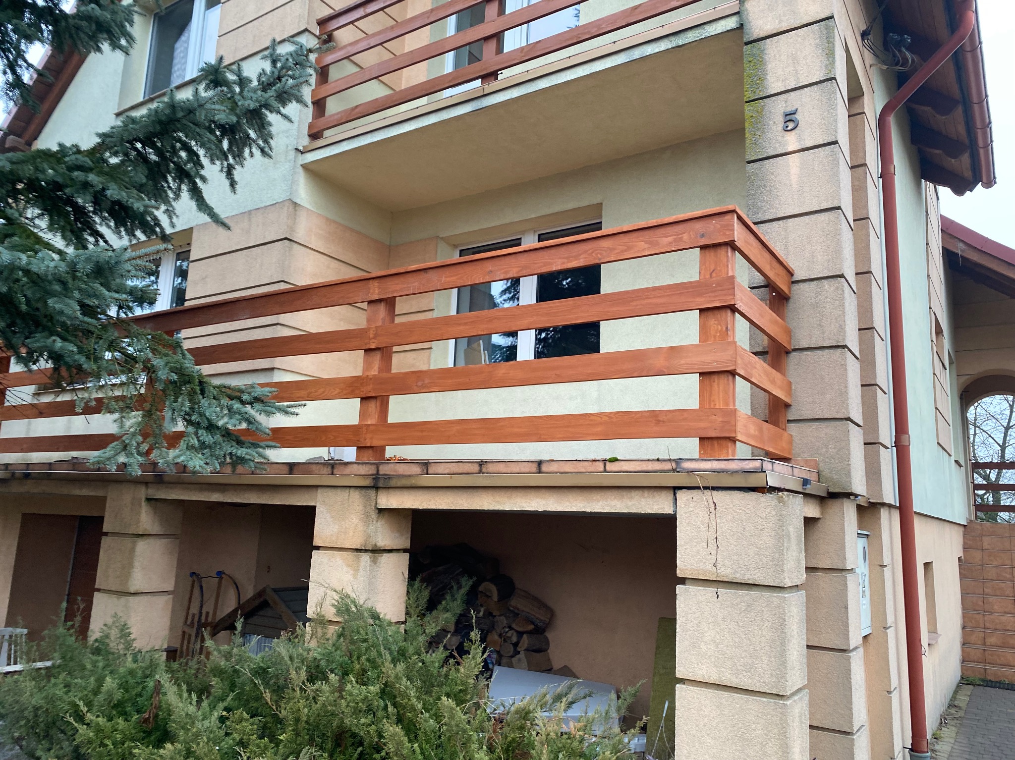 Dwupiętrowy dom z drewnianą balustradą balkonową, widok z poziomu gruntu, z widoczną numeracją budynku '5' i fragmentem iglastego drzewa po lewej stronie.
