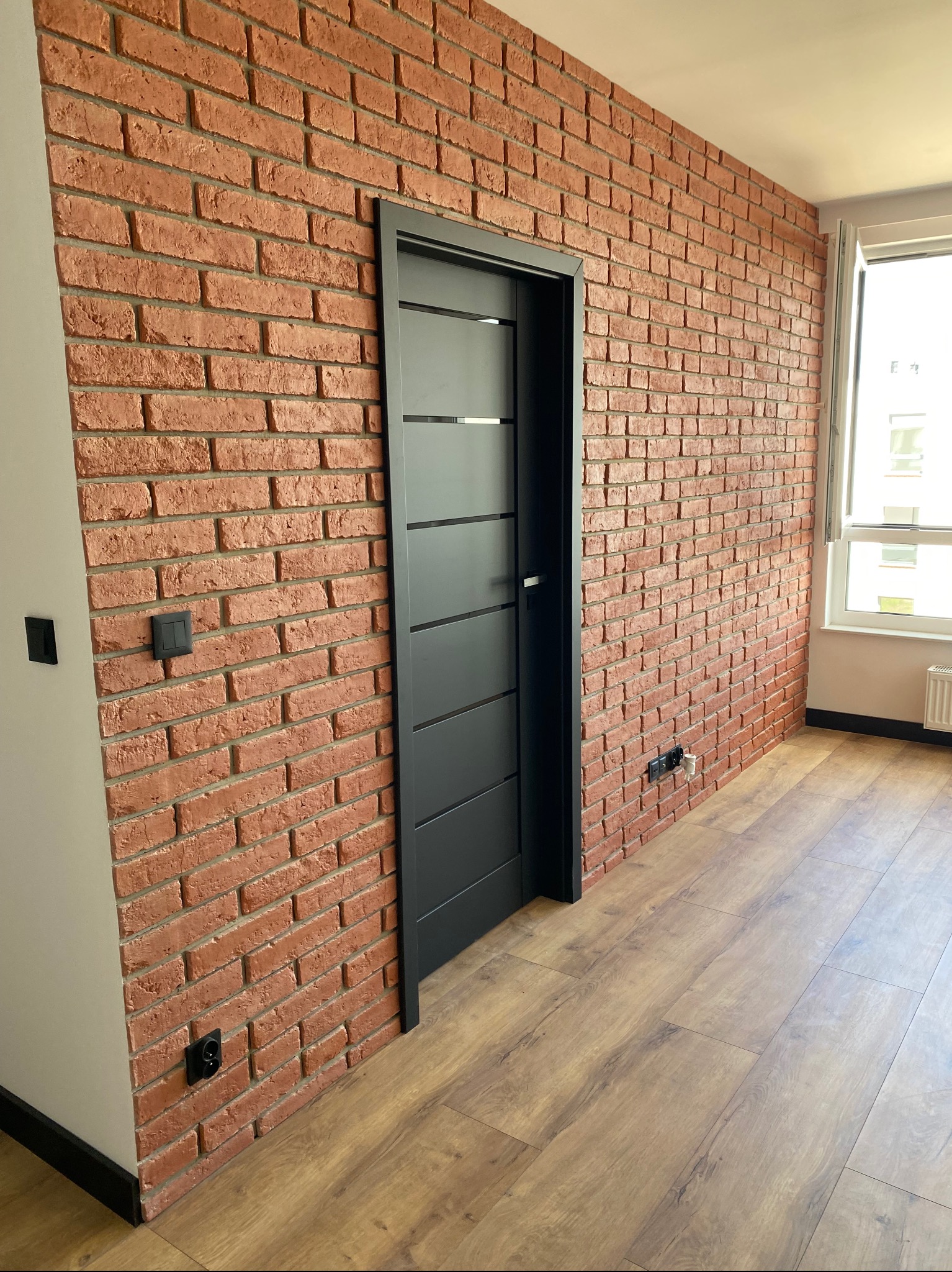 Wnętrze pokoju z dekoracyjną ścianą z czerwonej cegły, czarnymi drzwiami z poziomymi panelami i jasną drewnianą podłogą, widok na okno.