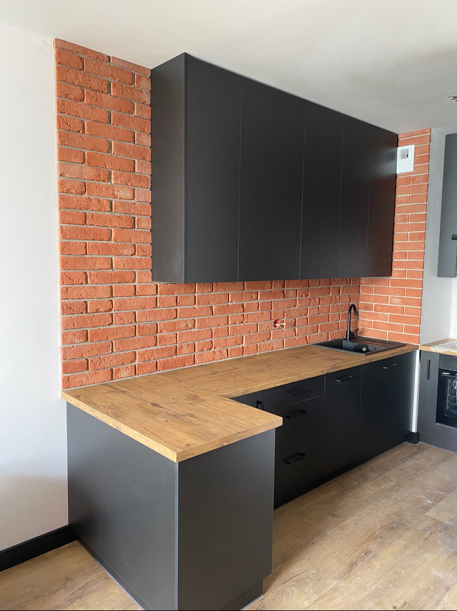 Nowoczesna kuchnia z czarnymi szafkami, drewnianym blatem i ceglaną ścianą, widoczna instalacja elektryczna nad blatem.