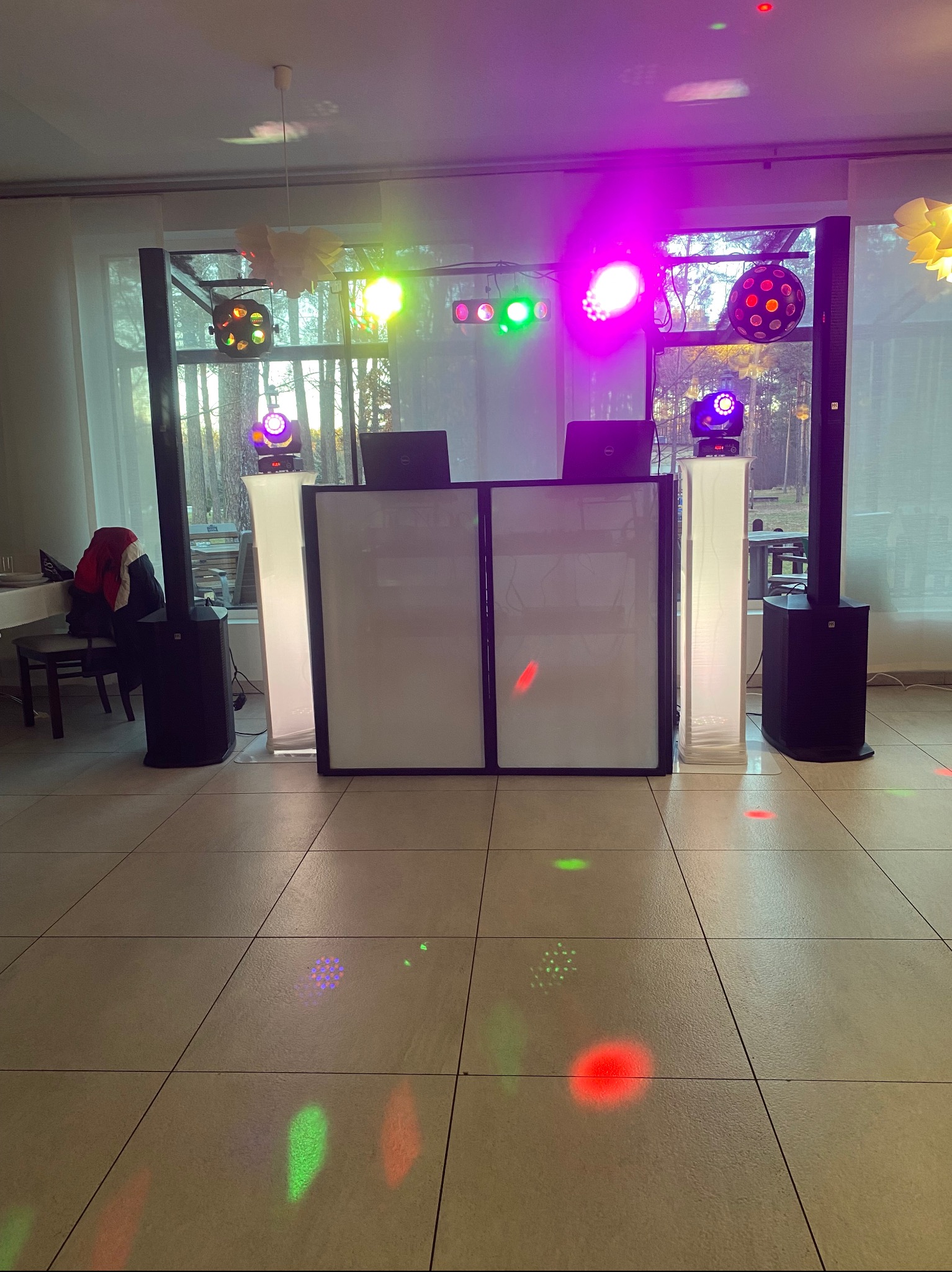 Rozstawiony sprzęt DJ-ski z oświetleniem scenicznym w sali. Widoczne kolumny, konsoleta DJ-ska z laptopami, reflektory i efekty świetlne na parkiecie.