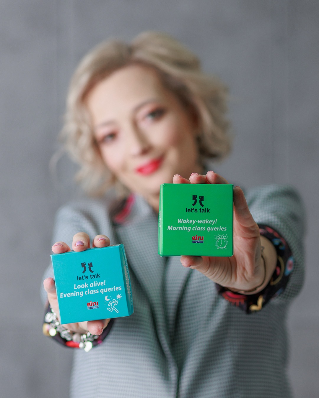 Kobieta prezentuje pudełka 'Let's talk' z hasłami 'Wakey-wakey! Morning class queries' i 'Look alive! Evening class queries' dla nauki angielskiego.