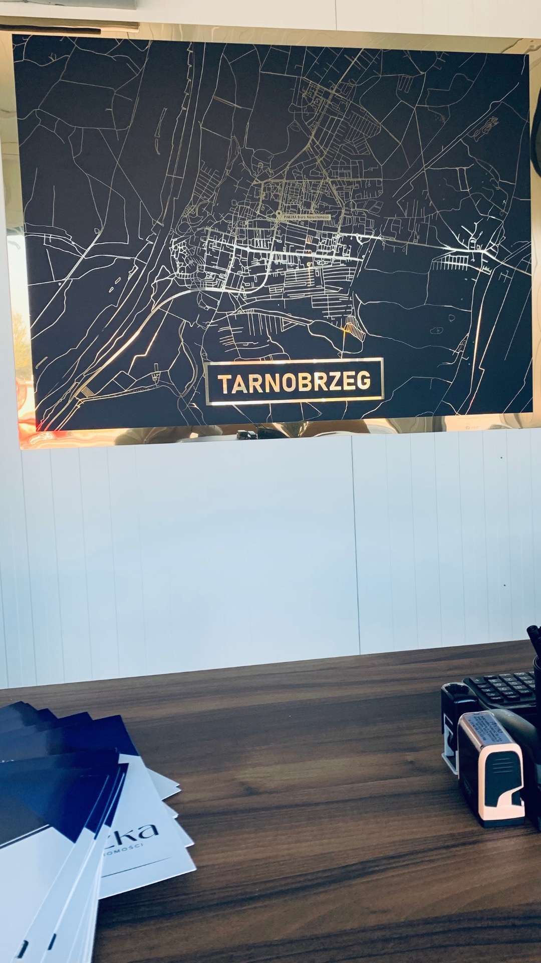 Mapa Tarnobrzega w złotej ramie na ścianie biura nieruchomości, na biurku ulotki i pieczątki.