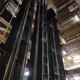 Wnętrze szybu windy z widoczną kabiną, linami nośnymi i konstrukcją stalową, oświetleniem technicznym i instalacjami pod stropem.