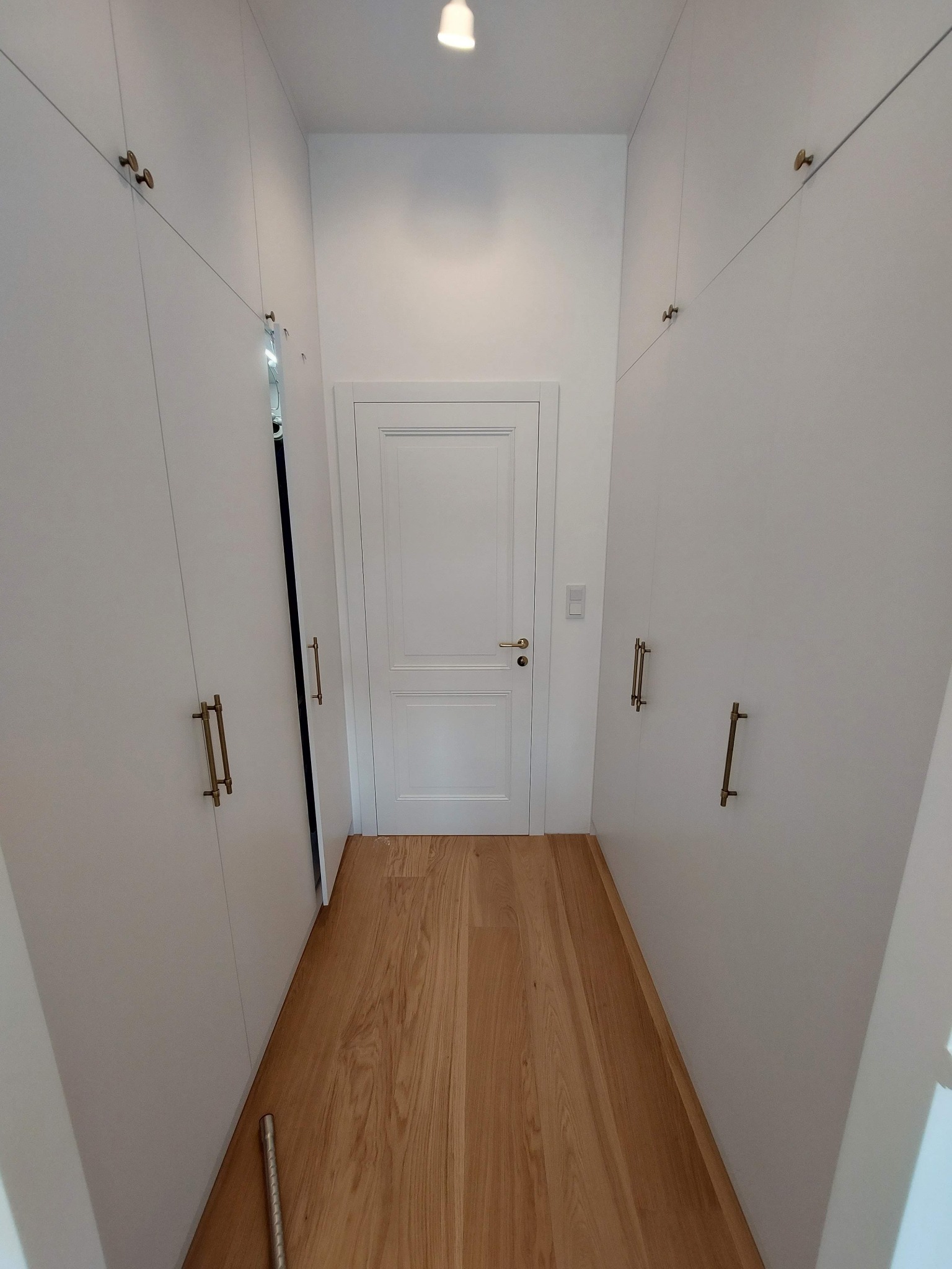 Białe szafy wnękowe z złotymi uchwytami, drewniana podłoga i białe drzwi w wąskim korytarzu. Minimalistyczny design, oświetlenie sufitowe.