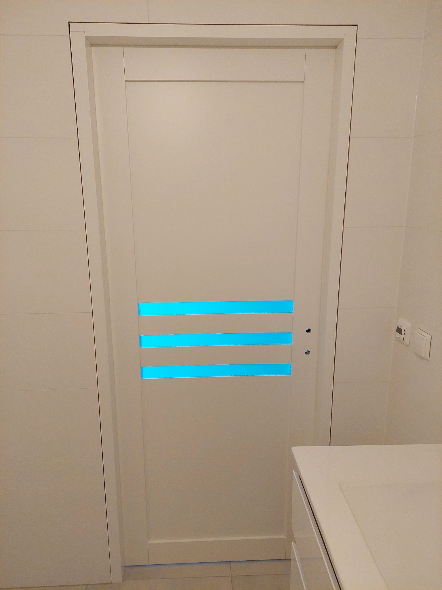 Białe drzwi wewnętrzne z trzema poziomymi, podświetlanymi na niebiesko paskami, zamontowane w łazience z białymi płytkami. Widoczny fragment szafki.