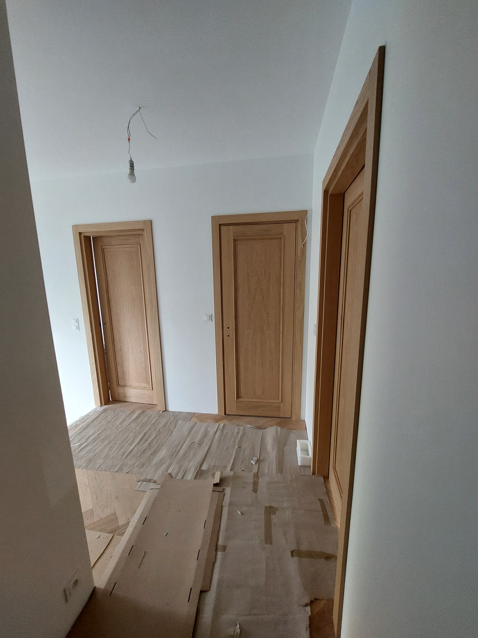 Wnętrze z trzema drewnianymi drzwiami po montażu, zabezpieczone podłoże kartonem i folią, surowy żyrandol. Widok z perspektywy korytarza.