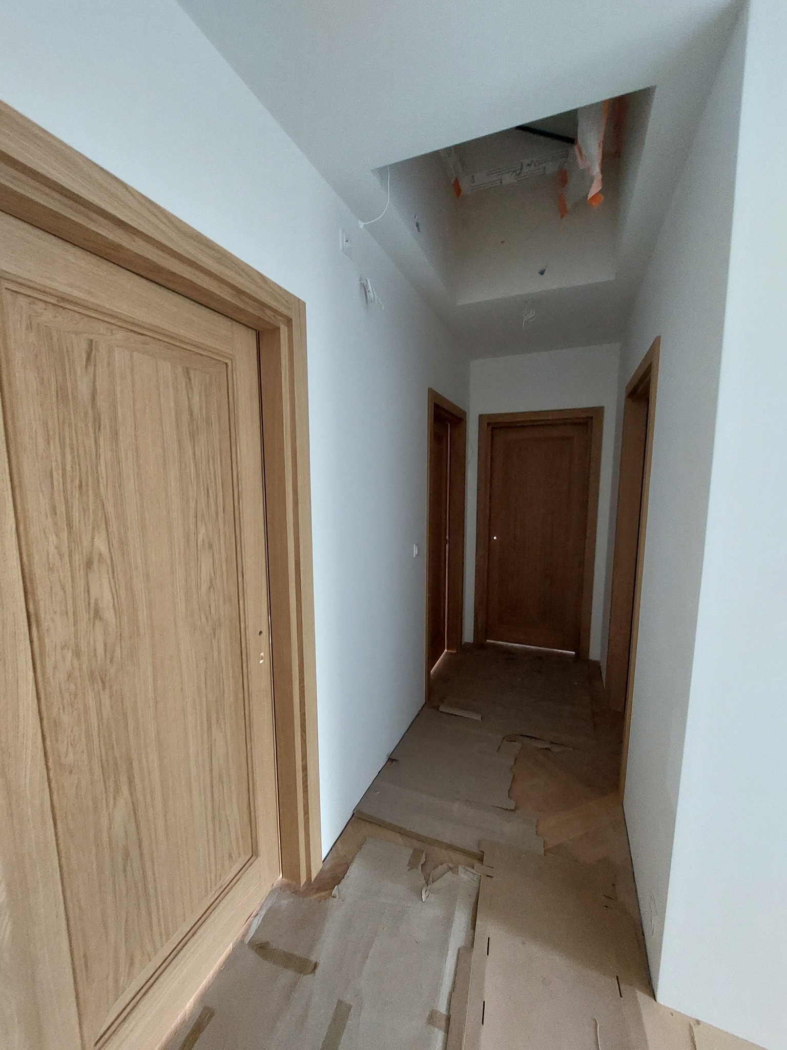 Jasne, drewniane drzwi wewnętrzne z ościeżnicą, widoczne w korytarzu w trakcie remontu. Podłoga zabezpieczona kartonem, sufit z otworem na oświetlenie.