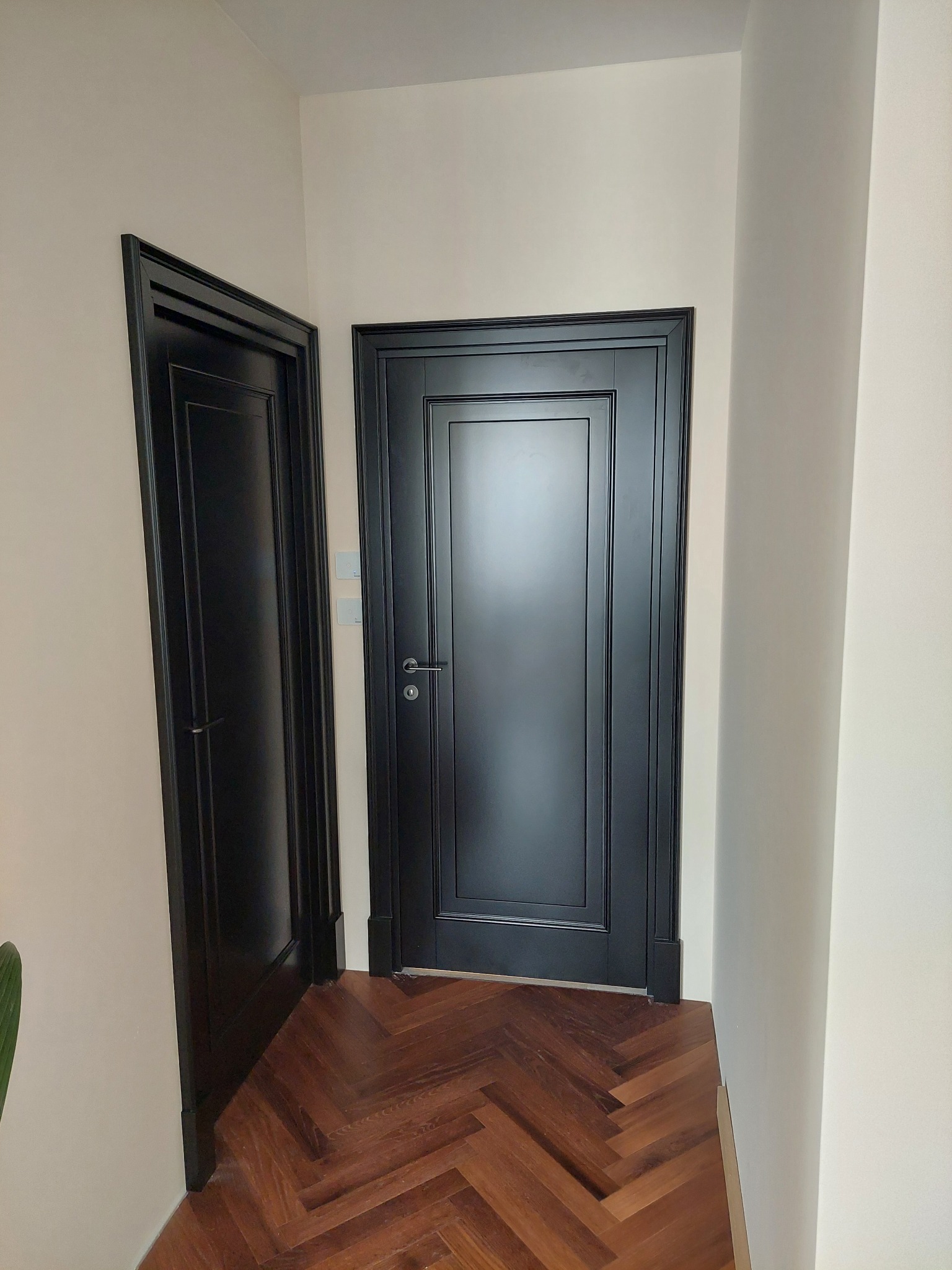 Dwa czarne, klasyczne drzwi wewnętrzne z frezowanymi ramkami, zamontowane w jasnym wnętrzu z parkietem ułożonym w jodełkę. Jeden otwarty, drugi zamknięty.