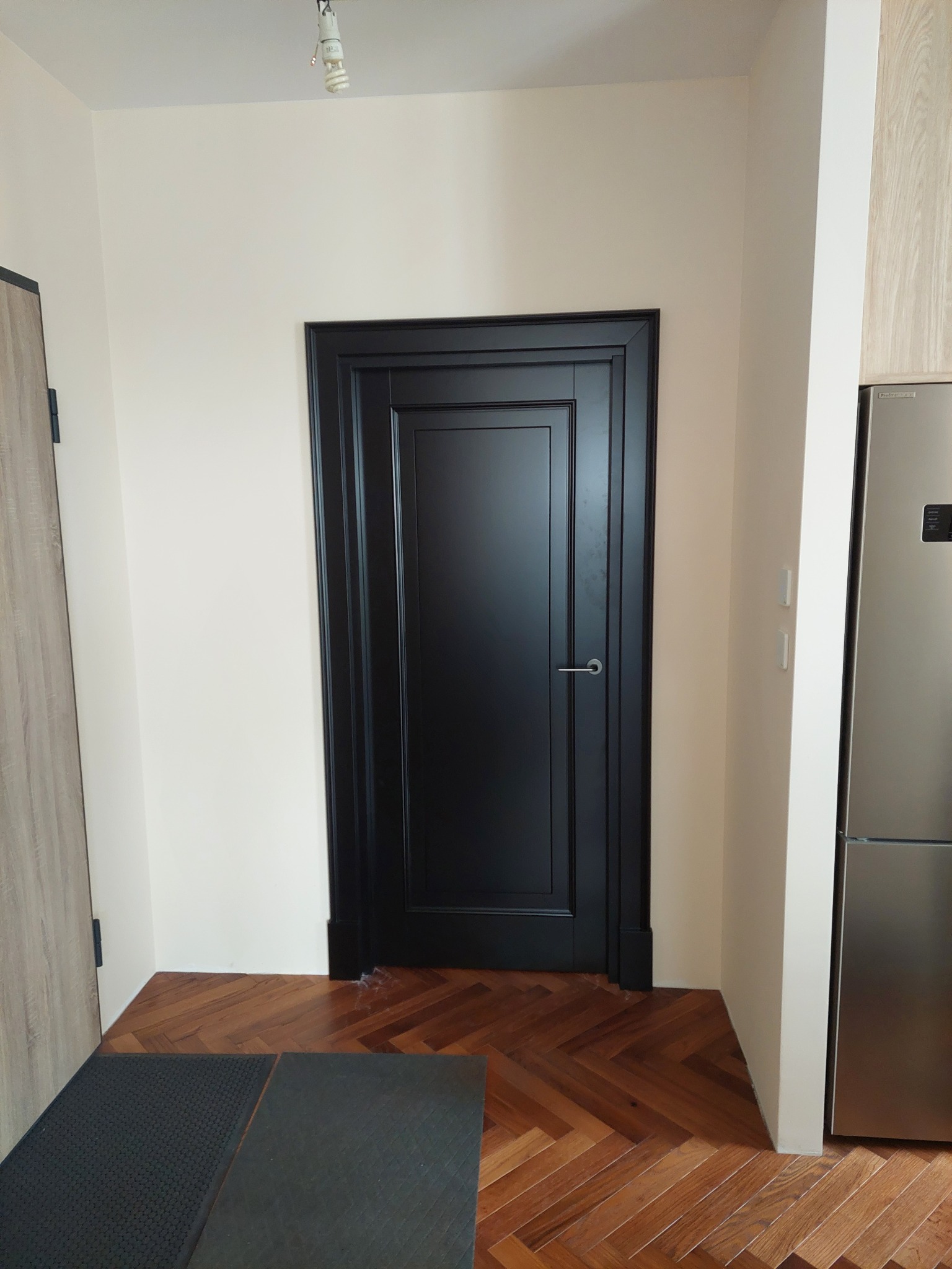 Eleganckie, czarne drzwi wewnętrzne z frezowanymi panelami, zamontowane w jasnym wnętrzu z parkietem ułożonym w jodełkę. Widoczny fragment lodówki i drzwi po lewej.