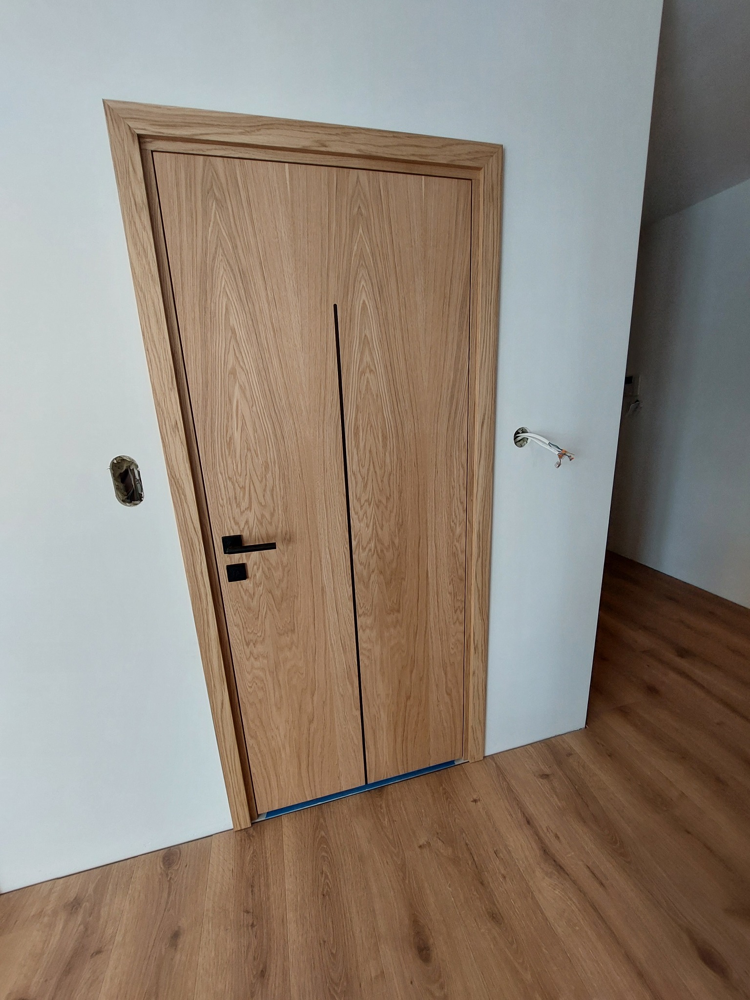 Zamknięte, drewniane drzwi wewnętrzne z czarną klamką, zamontowane w białej ścianie. Widoczne wykończenie podłogi panelami. Otwór po gniazdku elektrycznym.