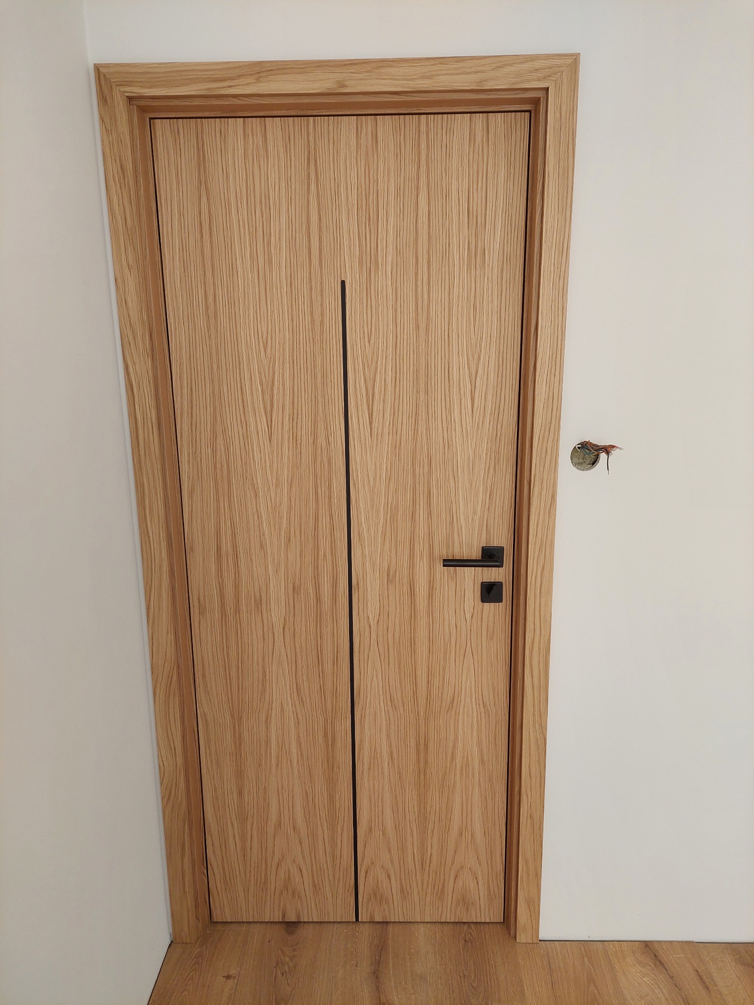 Zamknięte, drewniane drzwi z czarną klamką i pionowym, czarnym paskiem, w jasnej ościeżnicy. Na ścianie widoczne wystające kable.