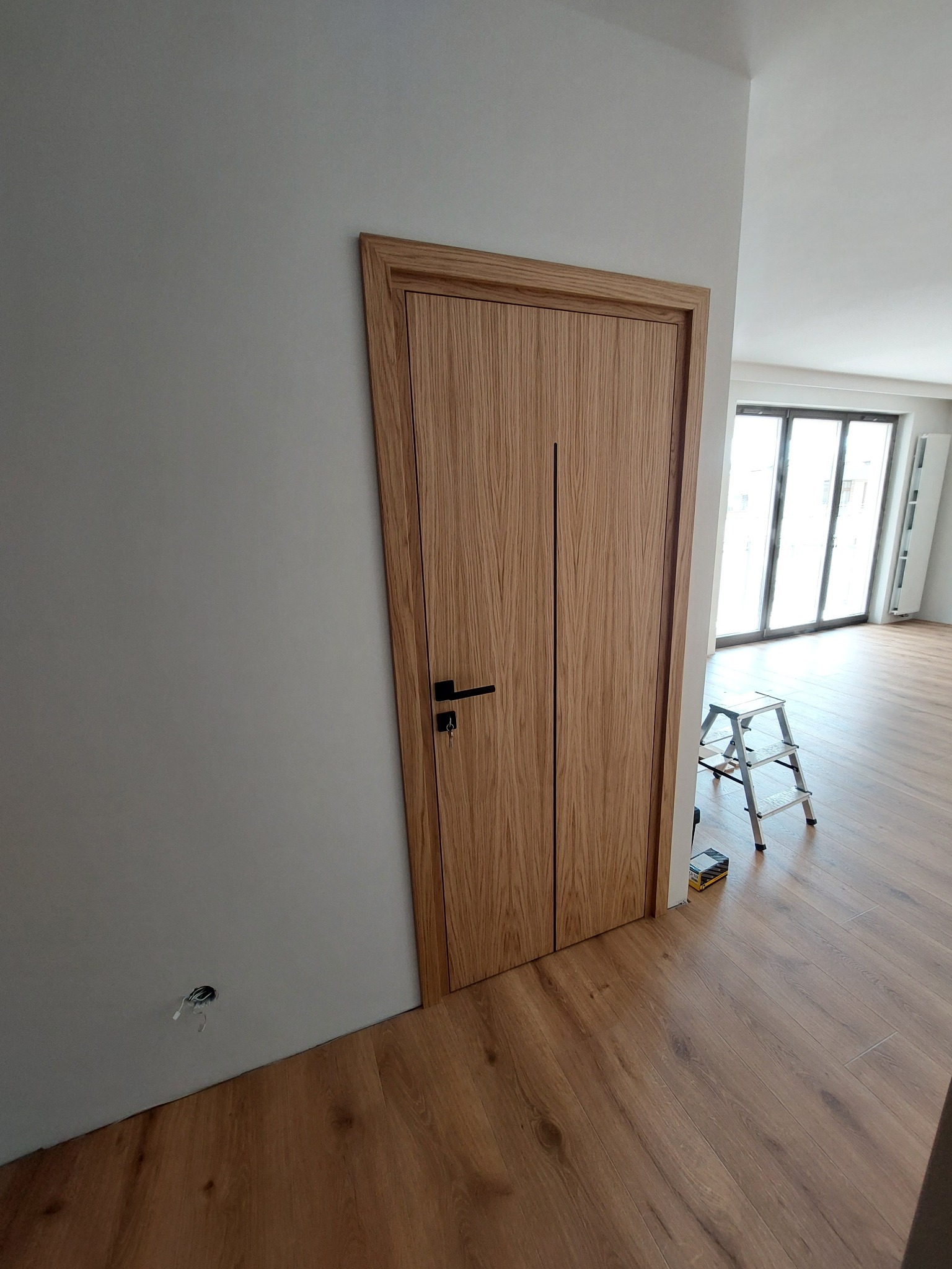Zamknięte, drewniane drzwi wewnętrzne z czarną klamką, widoczne na tle jasnej ściany i podłogi z paneli. W tle drabina malarska i okno balkonowe.