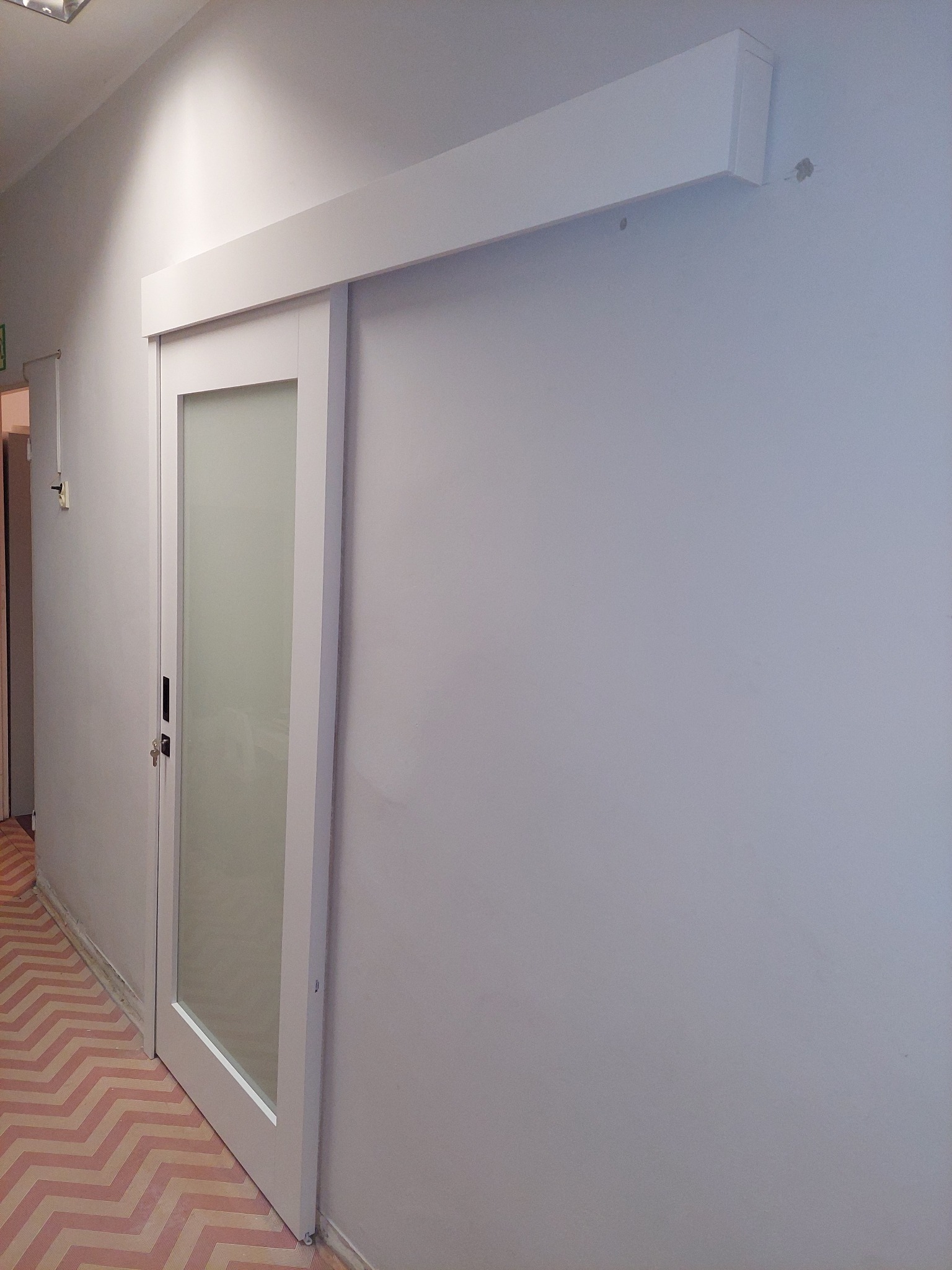 Białe, przesuwne drzwi z matową szybą, zamontowane w jasnym pomieszczeniu. Widoczna szyna nad drzwiami i wzorzysta podłoga w odcieniach różu i beżu.