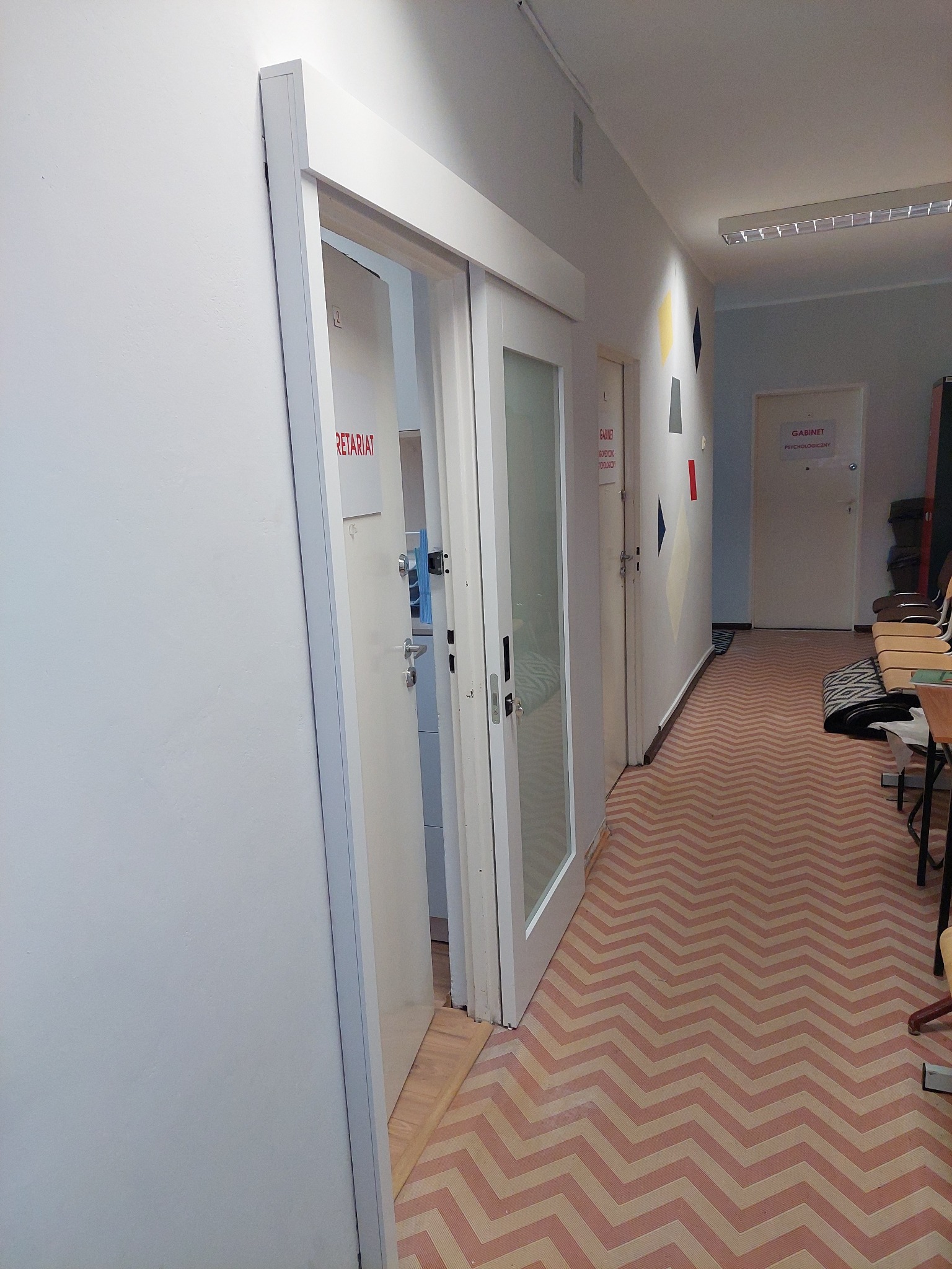 Białe drzwi z przeszkleniem w korytarzu biurowym, widok na wnętrze pomieszczenia z napisem 'Sekretariat'. Podłoga w zygzakowaty wzór. Oświetlenie sufitowe.