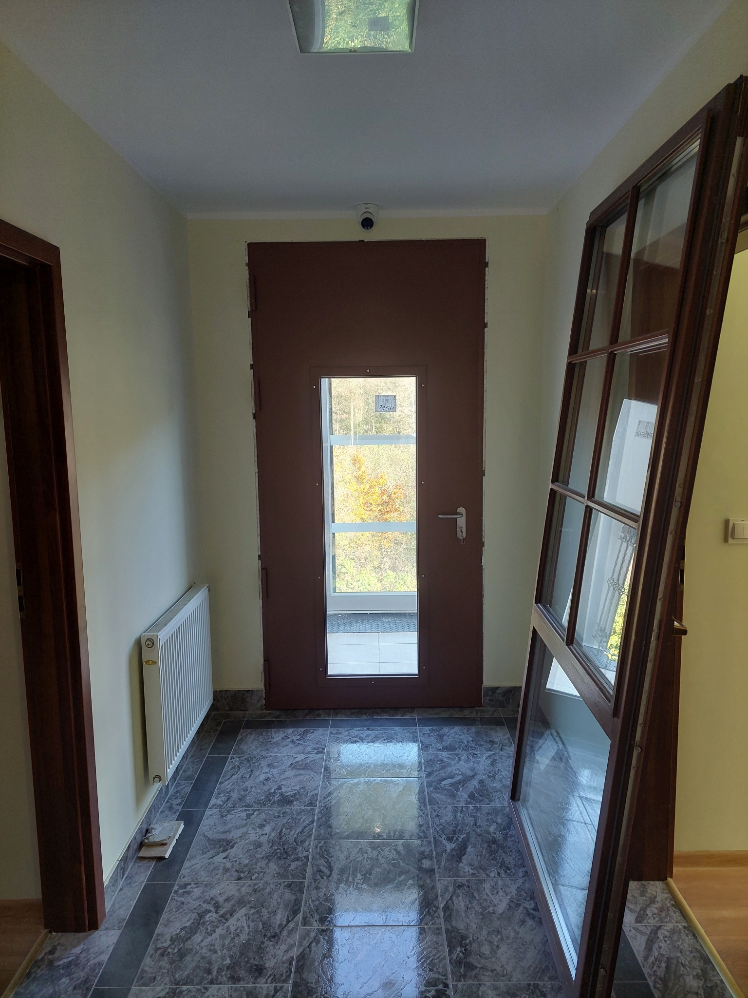 Korytarz z brązowymi drzwiami wejściowymi z szybą i oknem z drewnianą ramą opartym o ścianę. Widoczny grzejnik i szara marmurowa podłoga.