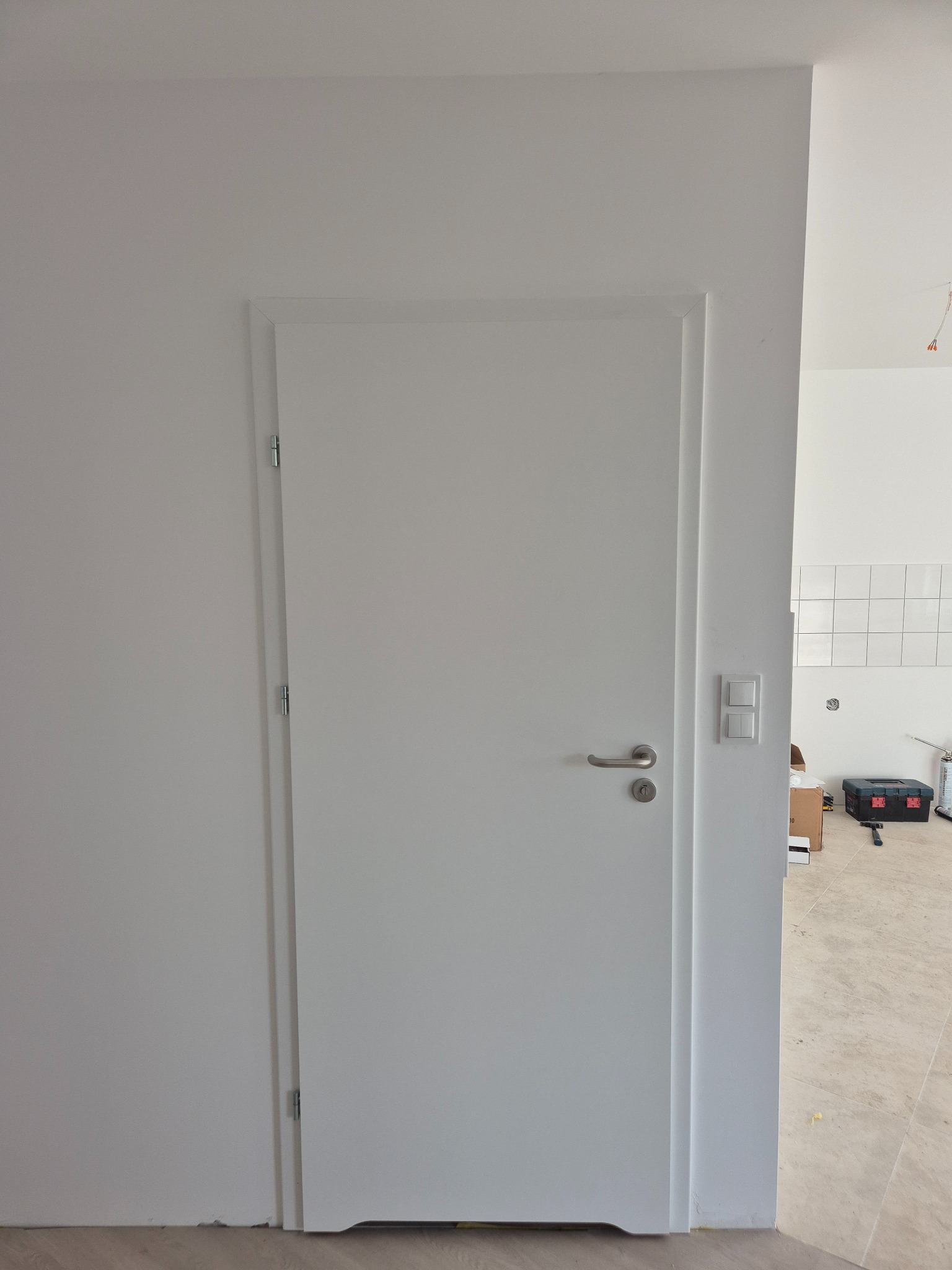 Białe, minimalistyczne drzwi wewnętrzne z srebrną klamką, zamontowane w białej ścianie. W tle widoczne narzędzia i płytki, sugerujące świeżo wykończone wnętrze.