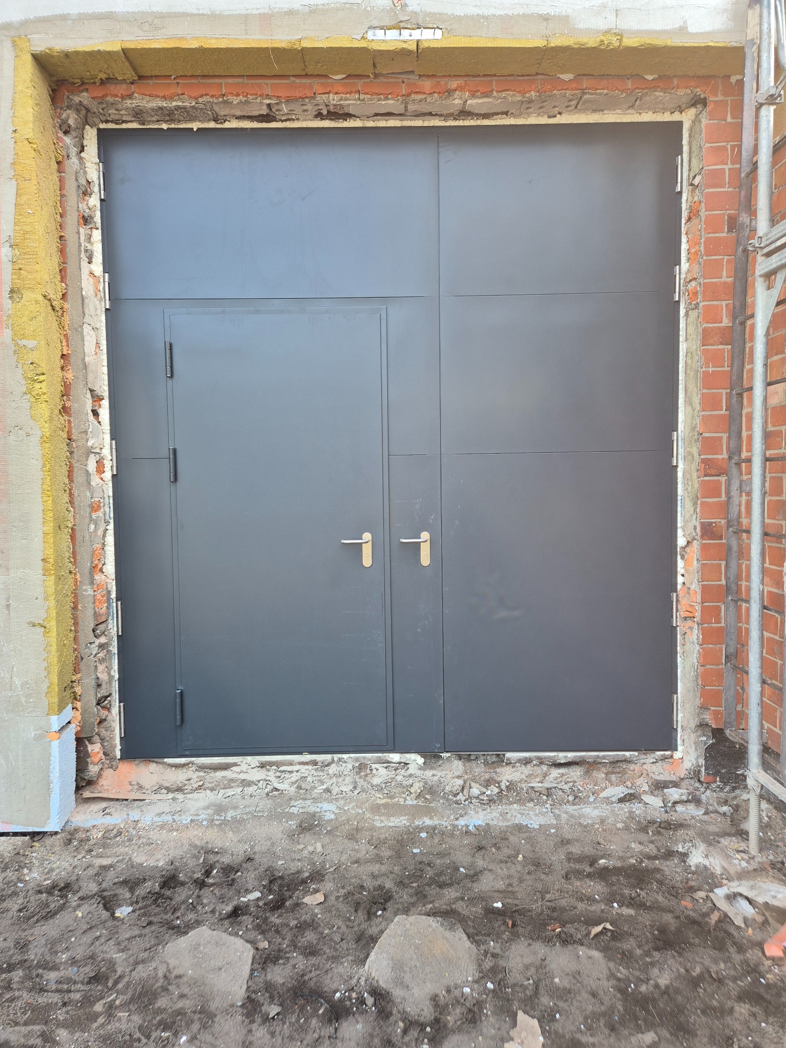 Szare, dwuskrzydłowe drzwi stalowe z ościeżnicą w trakcie montażu w surowym otworze muru z widoczną izolacją termiczną i rusztowaniem z boku.