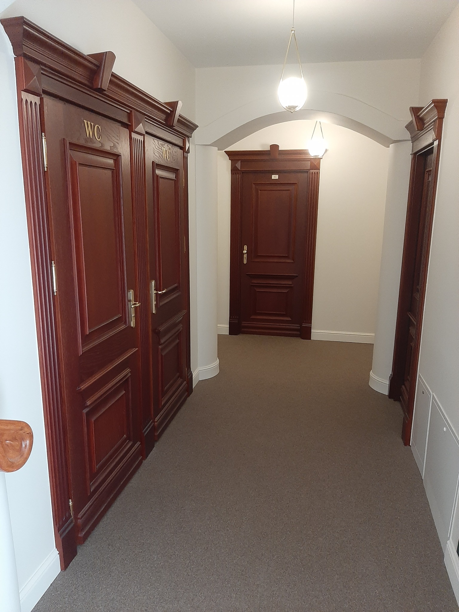 Korytarz z trzema parami ciemnobrązowych, drewnianych drzwi z ozdobnymi framugami. Dwoje drzwi oznaczonych 'WC'. W tle widoczne kolejne drzwi pod łukiem.