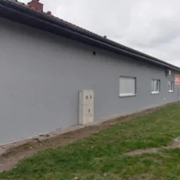 Szara elewacja budynku z widocznymi oknami, rynnami, skrzynką elektryczną i fragmentem trawnika. Budynek ma brązowy dach.