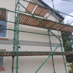 Elewacja domu w trakcie prac wykończeniowych, widoczne rusztowanie, okna zabezpieczone folią, świeżo pomalowana ściana z szarym pasem.