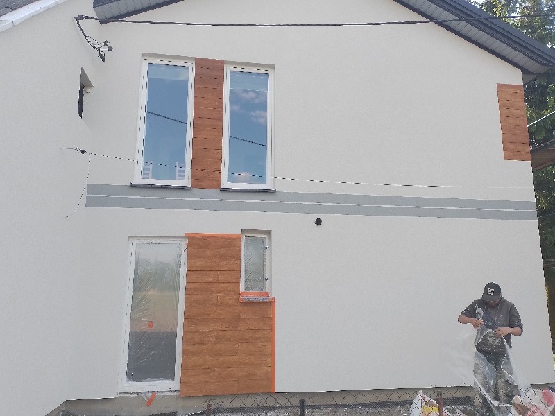 Elewacja domu jednorodzinnego z fragmentami imitacji drewna, zabezpieczonymi oknami i drzwiami folią malarską, przygotowywana do malowania przez pracownika w czapce.