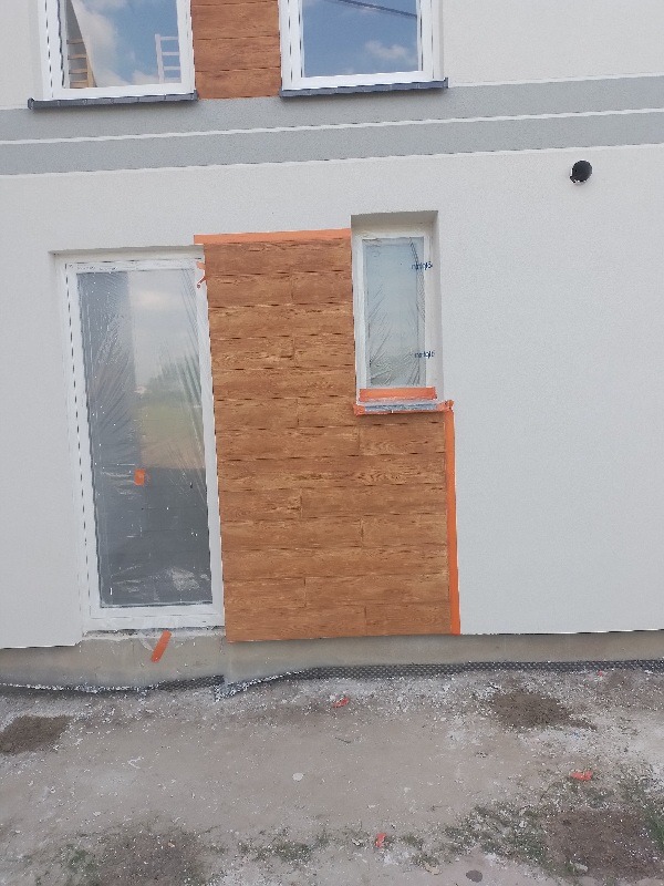 Montaż paneli imitujących drewno na elewacji budynku, widoczne okna zabezpieczone folią, prace w toku.