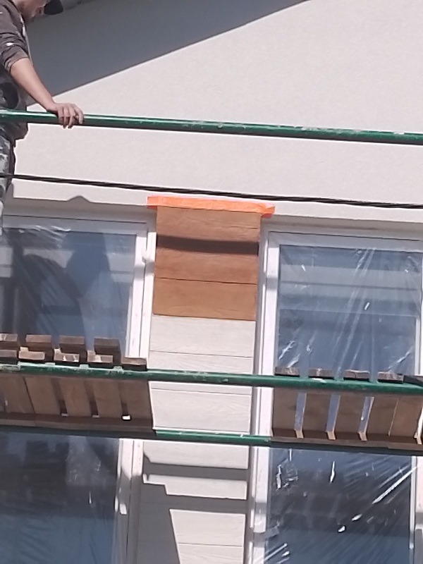 Prace elewacyjne: fragment ściany z nałożonym tynkiem imitującym drewno, zabezpieczone okna folią, widoczne rusztowanie.