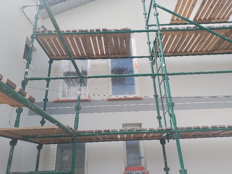 Elewacja budynku w trakcie prac wykończeniowych, widoczne rusztowanie, okna zabezpieczone folią, szary pas na ścianie.