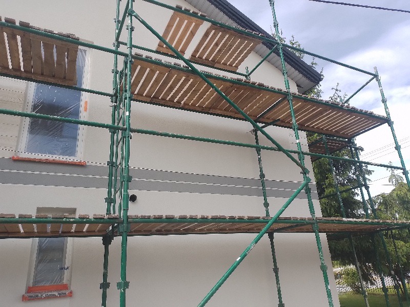 Elewacja domu w trakcie prac wykończeniowych, widoczne rusztowanie, okna zabezpieczone folią, świeżo pomalowana ściana z szarym pasem.