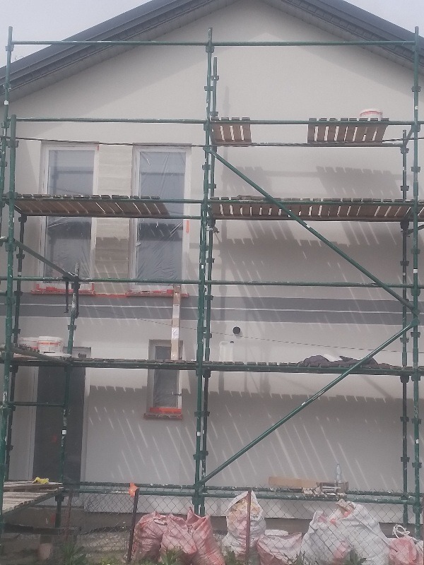 Elewacja domu w trakcie prac wykończeniowych, widoczne rusztowania, okna zabezpieczone folią, worki z materiałami budowlanymi u podnóża ściany, świeżo nałożony tynk.