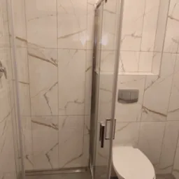 Nowoczesna łazienka z kabiną prysznicową ze szklanymi drzwiami, białe płytki imitujące marmur z brązowymi żyłami, chromowana bateria, podwieszana toaleta z szarym przyciskiem spłukującym i uchwyt...