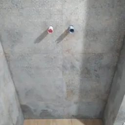 Wykończona ściana łazienki z dekoracyjnymi płytkami w odcieniach szarości i beżu, widoczne przyłącza wodne i odpływ liniowy w podłodze z płytek imitujących drewno.