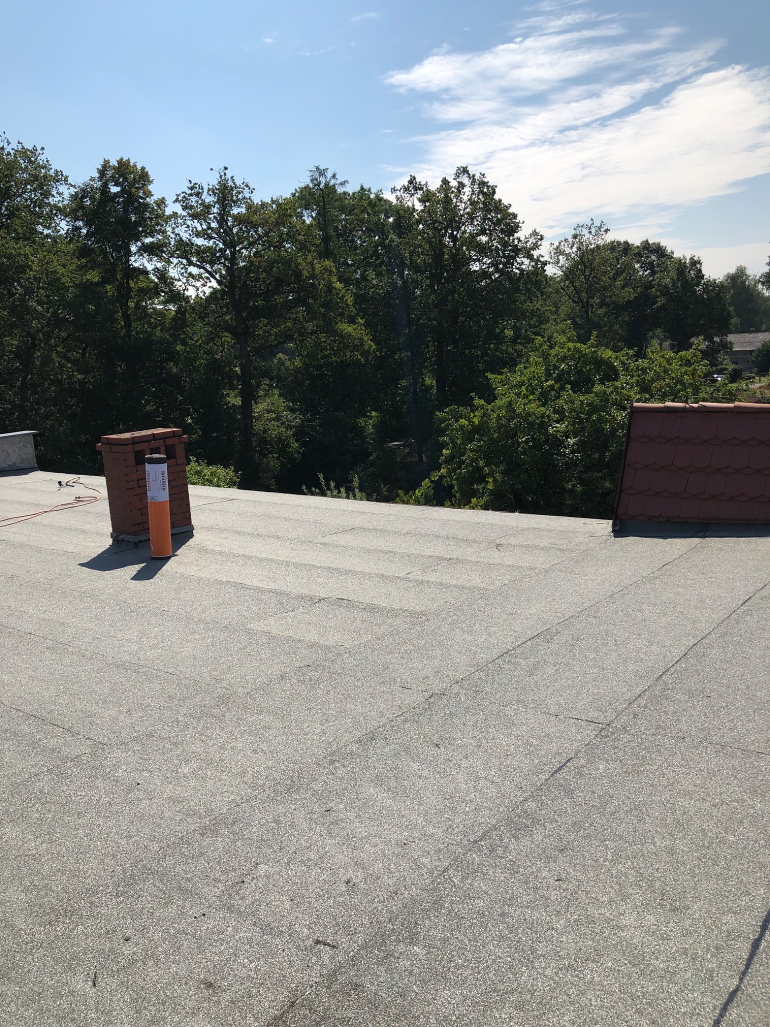 Płaski dach pokryty papą termozgrzewalną w kolorze szarym, z widocznym kominem z czerwonej cegły i tubą z materiałem dekarskim, w tle drzewa i błękitne niebo z chmurami.