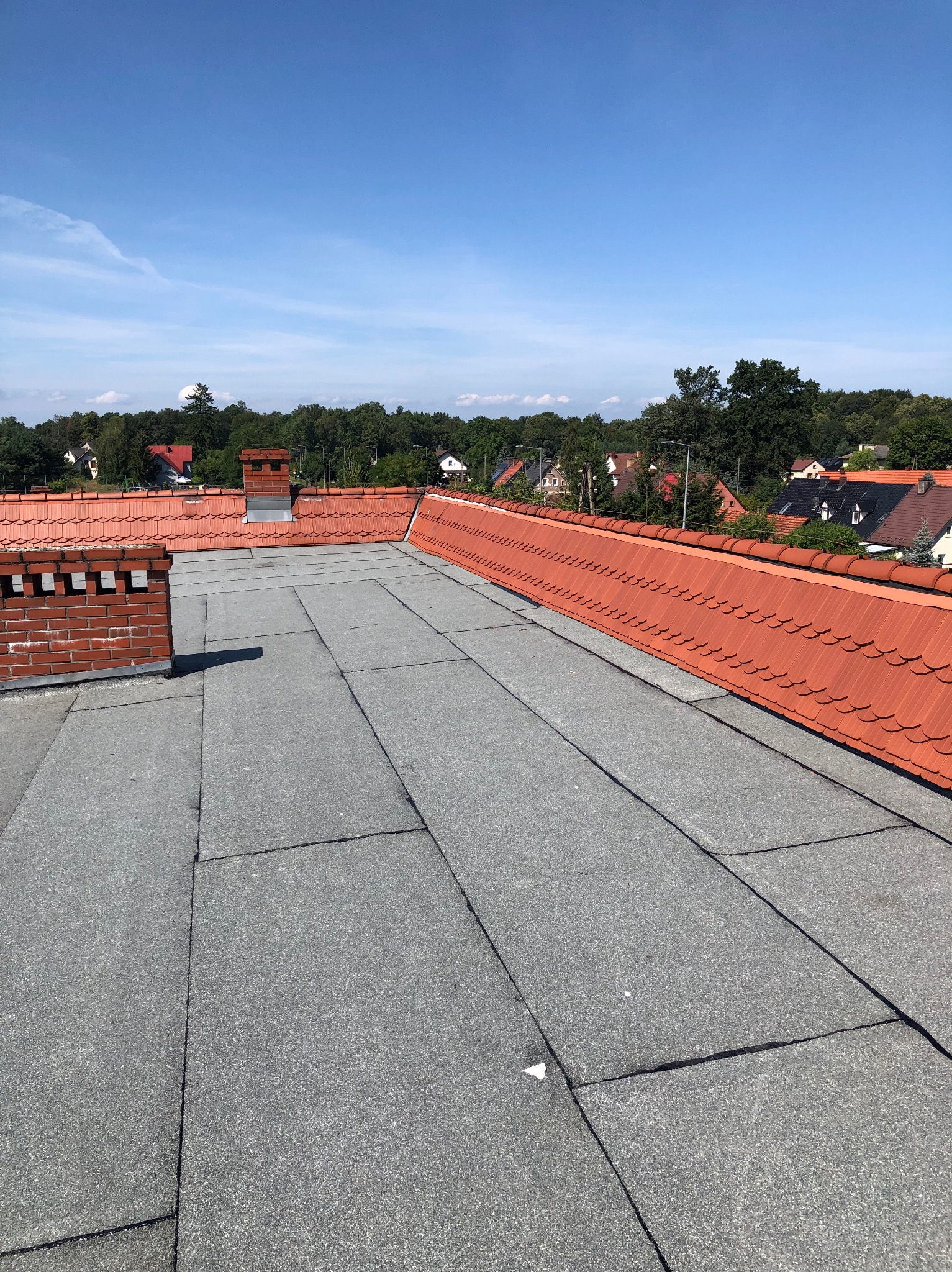 Nowe pokrycie dachu papą termozgrzewalną w kolorze szarym, widoczne kominy z cegły i dachówka ceramiczna na krawędzi dachu, w tle zabudowa jednorodzinna i zieleń.