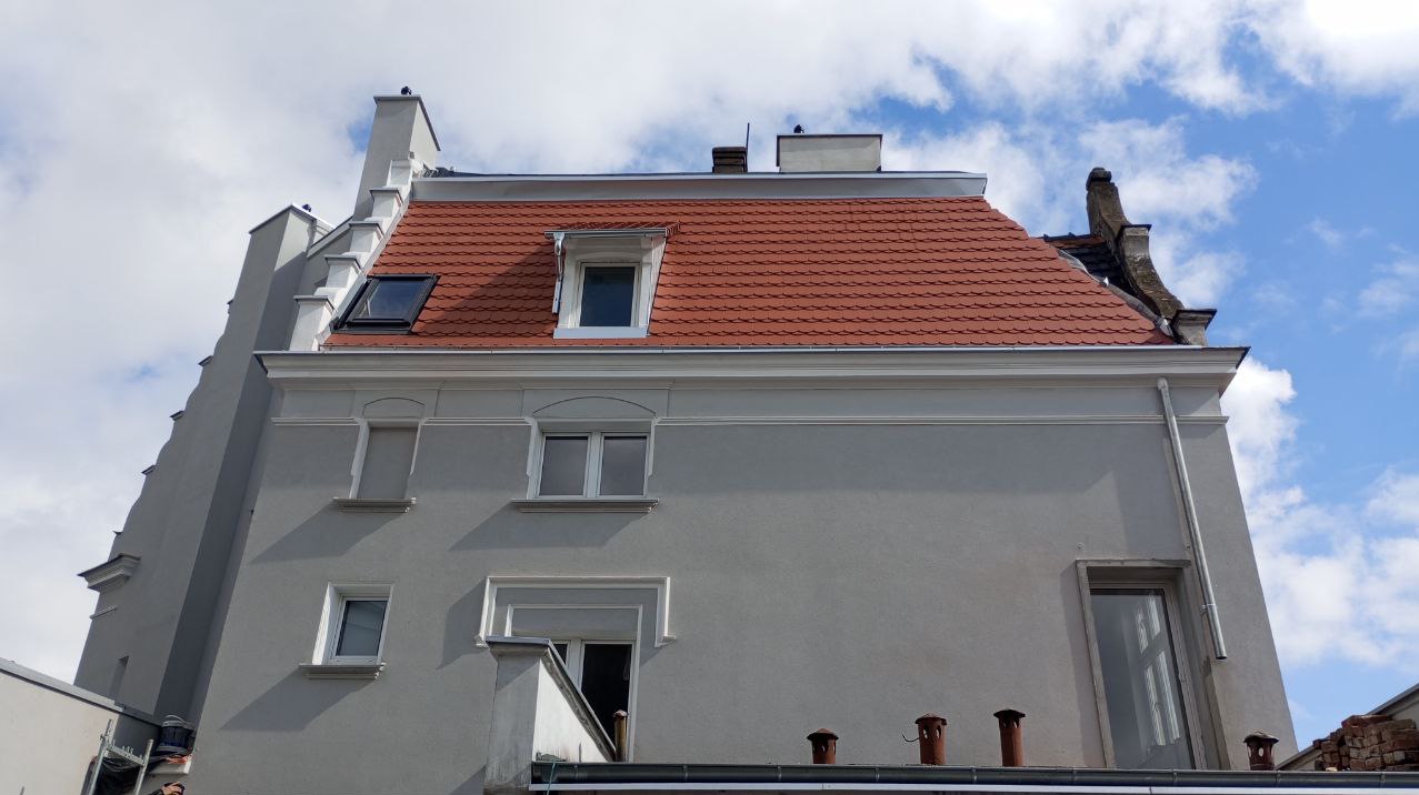 Budynek z nową, czerwoną dachówką karpiówką, widoczne okna dachowe, kominy i ozdobne elementy architektoniczne, na tle błękitnego nieba z chmurami.