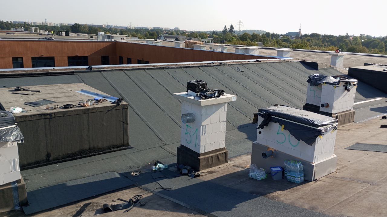 Widok z góry na płaski dach pokryty papą termozgrzewalną, z kominami wentylacyjnymi z betonu komórkowego oznaczonymi numerami 50 i 51, częściowo zabezpieczonymi folią. W tle budynki i zieleń.