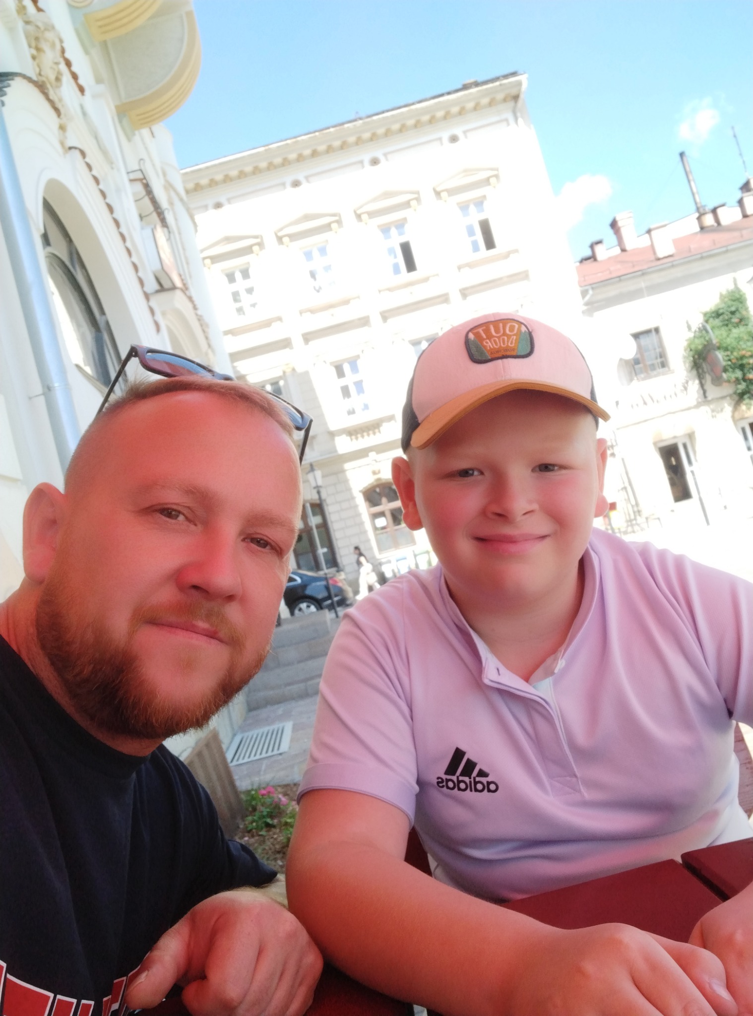 Selfie mężczyzny z brodą i chłopca w czapce na tle budynku z jasną elewacją i ozdobnymi detalami architektonicznymi.