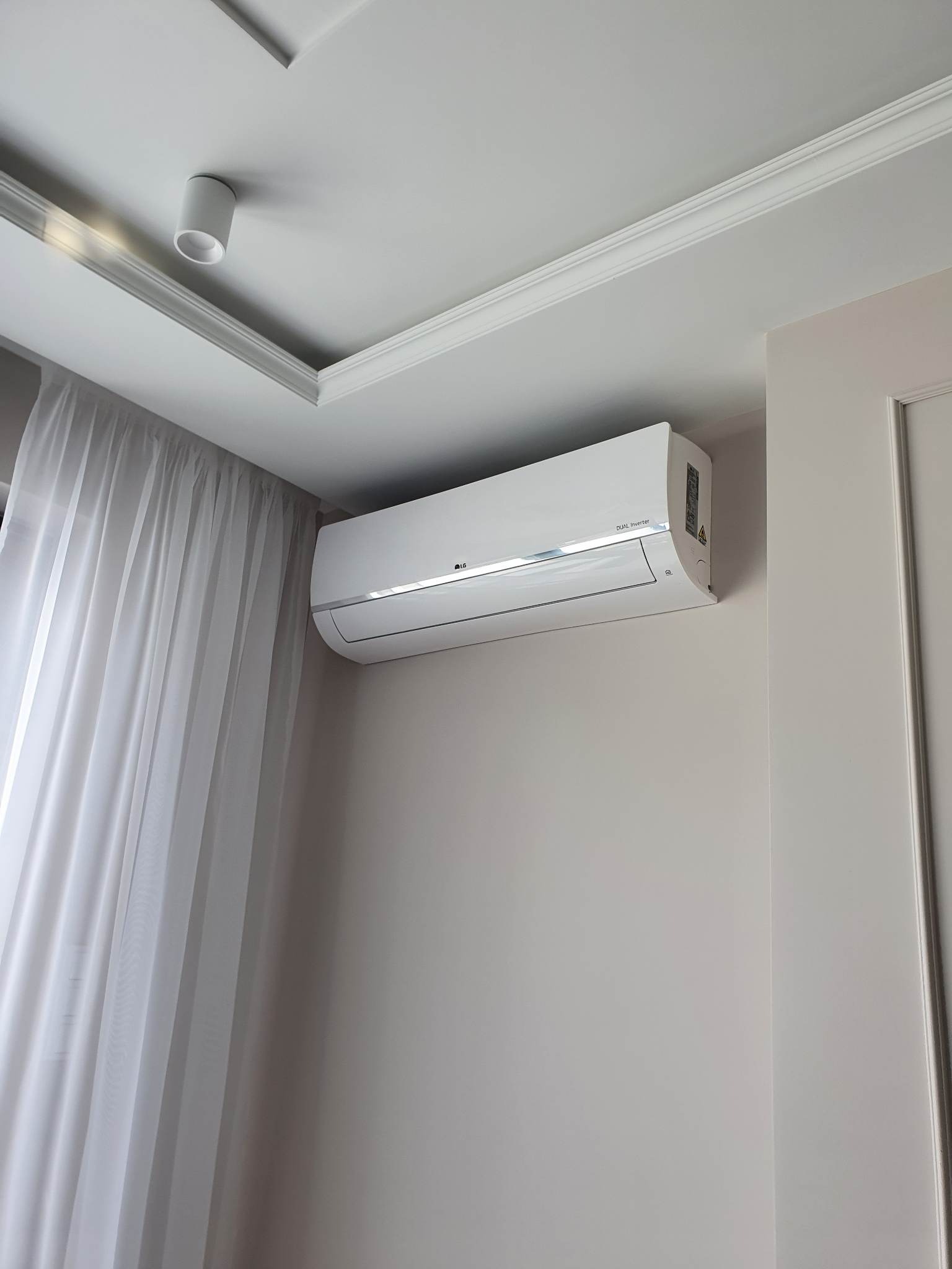 Biały klimatyzator LG Dual Inverter zamontowany na ścianie w jasnym pomieszczeniu z delikatnymi, białymi zasłonami i ozdobnym oświetleniem sufitowym.