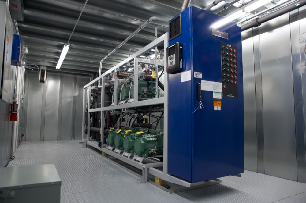 Kompleksowa instalacja chłodnicza z rzędami sprężarek w metalowej obudowie, widoczne przewody, panel sterowania i oświetlenie przemysłowe w pomieszczeniu technicznym.