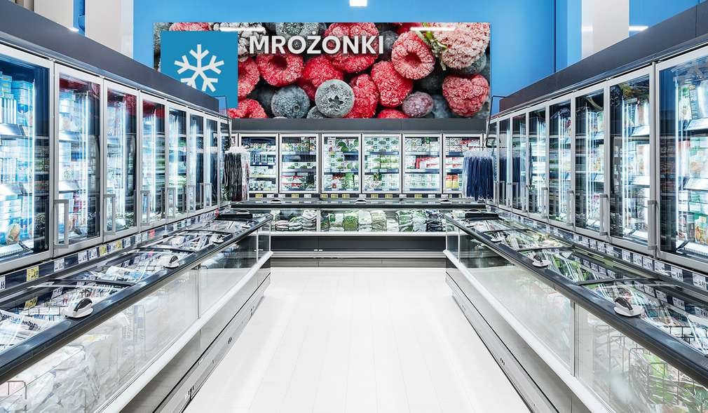 Długi rząd przeszklonych lodówek w supermarkecie z produktami mrożonymi, nad nimi baner z napisem 'MROŻONKI' i grafiką owoców pokrytych szronem.