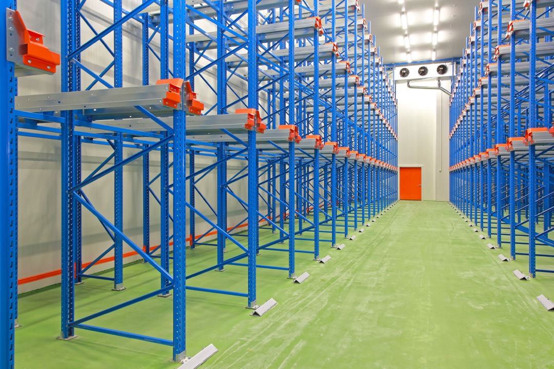 Puste, niebieskie regały magazynowe z pomarańczowymi elementami mocującymi w chłodni z zieloną podłogą i pomarańczowymi rurami na ścianie.