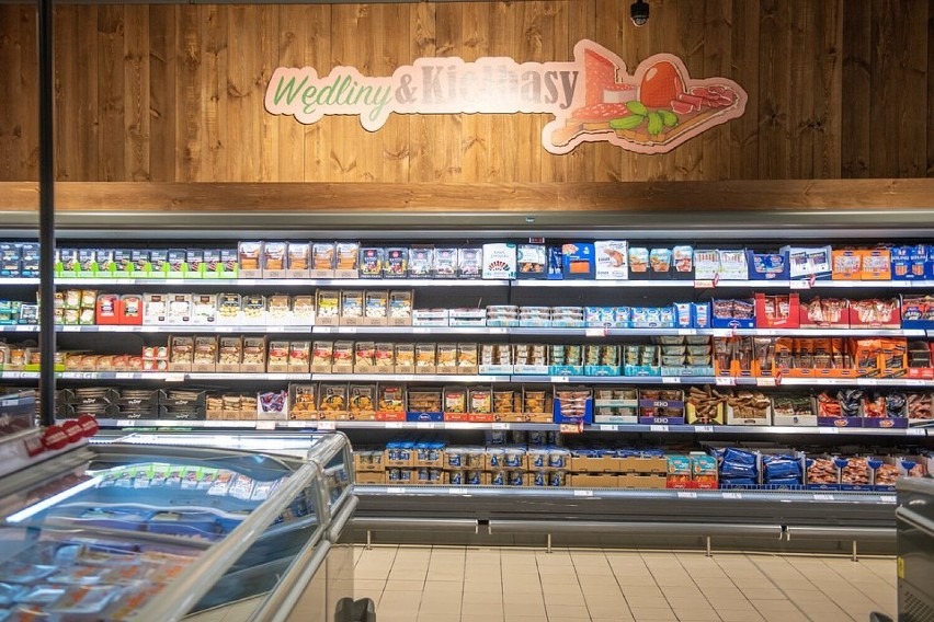 Półki chłodnicze w supermarkecie wypełnione różnorodnymi wędlinami i kiełbasami w opakowaniach, z drewnianym panelem i dekoracyjnym napisem 'Wędliny & Kiełbasy' nad nimi.
