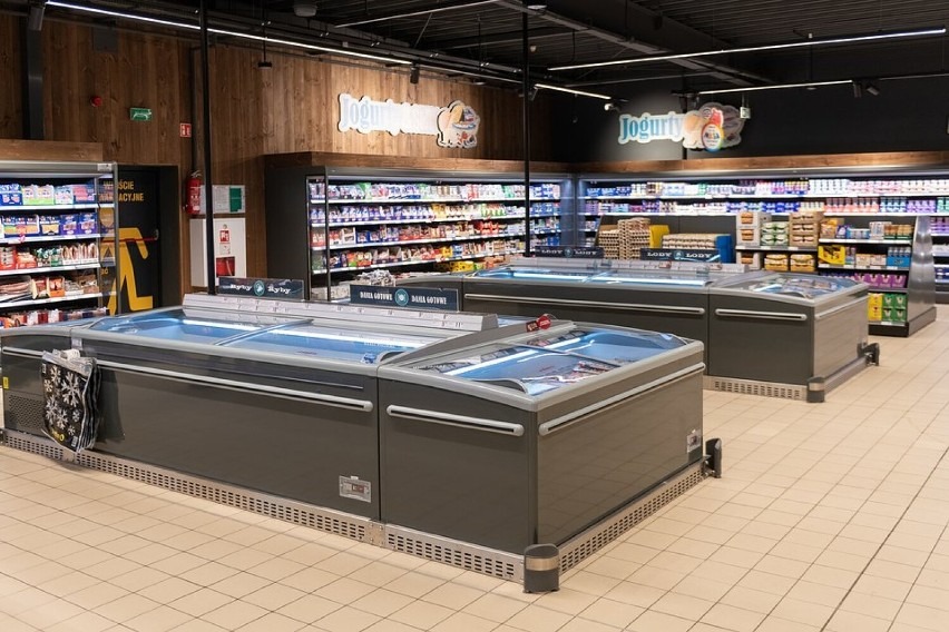 Wnętrze sklepu spożywczego z rzędami chłodziarek na lody i mrożonki, oświetlone lampami sufitowymi, z widocznymi półkami pełnymi produktów w tle.